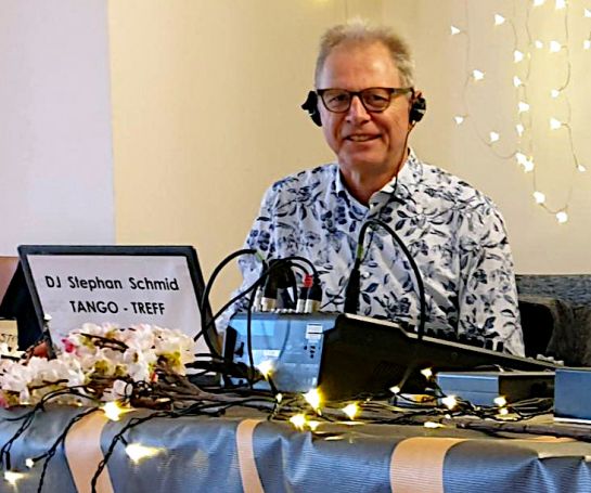 Lachender Mann mit Brille, Köpfhörern und grauen Haaren hinter einem Mischpult. Tisch und Hintergrund sind mit Blumen, Lichterketten und einer Tischdecke geschmückt