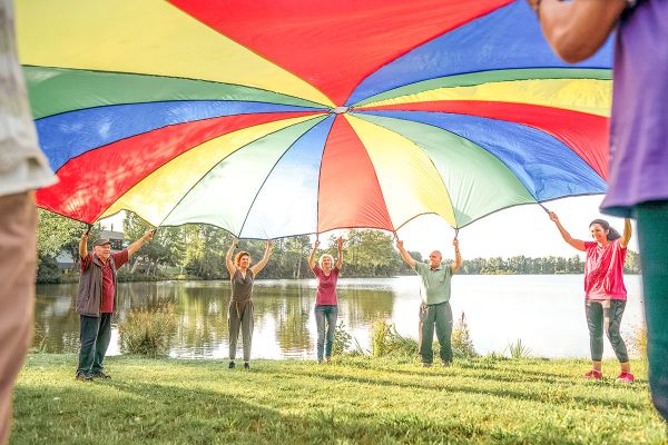 Fotografie - Männer und Frauen halten ein buntes Tuch über ihren Köpfen; im Hintergrund ist ein See zu sehen