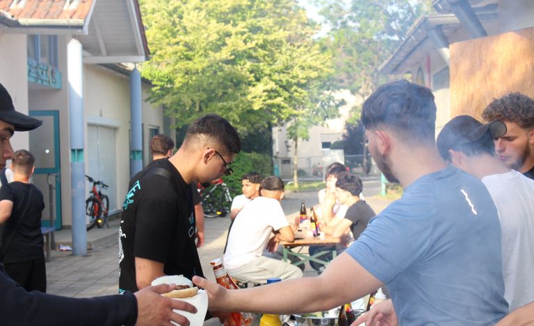 Eindrücke vom Sommerfest des Jugendzentrum Arena. Viele Jugendliche sind zu sehen. Im Vordergrund überreicht ein junger Mann einem anderen einen Burger und im Hintergrund sitzen mehrere junge Männer an einem Tisch und stoßen mit Spezi-Flaschen an