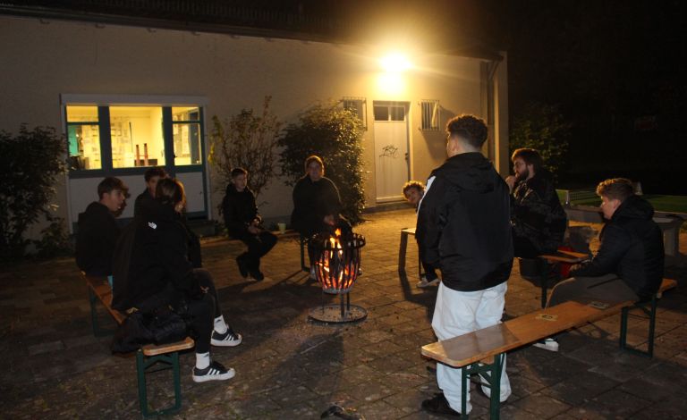 Das Bild zeigt eine Gesprächsrunde im Dunkeln auf dem Außengelände des Jugendzentrum. In der Mitte steht ein Feuerkorb um welchen in einem Viereck mehrere Bierbänke angeordnet sind, auf denen mehrere Menschen sitzen. Im Vordergrund des Bildes steht ein Kind, mit dem Rücken zu Kamera. Das Bild wird erhellt durch eine Außenleuchte am Haus.