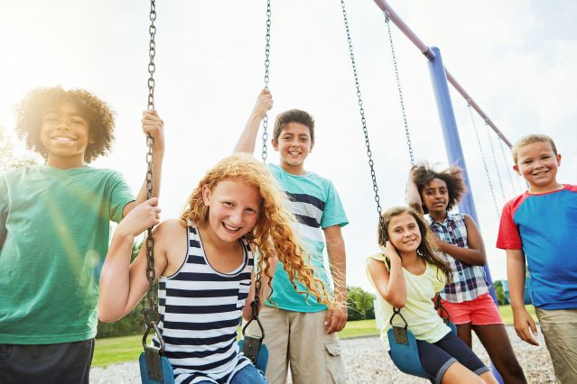 Das Bild zeigt eine diverse, lachende Kindergruppe. Ein Teil der Kinder sitzt auf Schaukeln.