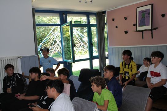 Das Bild wurde in einem Innenraum aufgenommen. Im Hintergrund sieht man ein Fenster. Im Vordergrund sind einige Kinder zu sehen, die gebannt nach links vorne gucken, wo sich der Fernseher befindet, auf dem gerade das Playstation-Spiel Fifa gespielt wird.