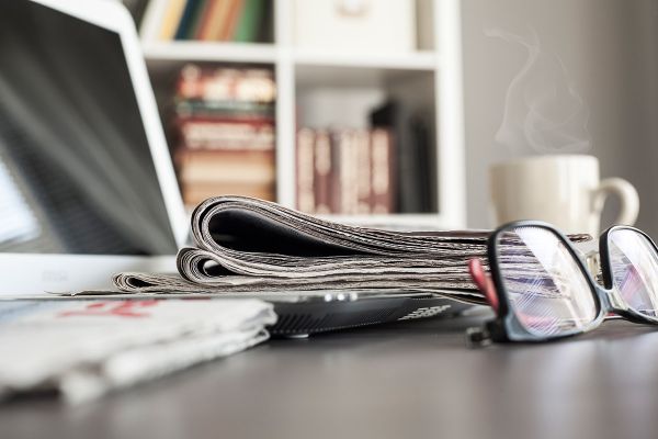 Symbolbild für Aktuelles - Bildschirm, Zeitung und Brille am Schreibtisch, im Hintergrund Kaffeetasse