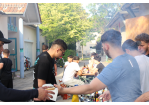 Eindrücke vom Sommerfest des Jugendzentrum Arena. Viele Jugendliche sind zu sehen. Im Vordergrund überreicht ein junger Mann einem anderen einen Burger und im Hintergrund sitzen mehrere junge Männer an einem Tisch und stoßen mit Spezi-Flaschen an