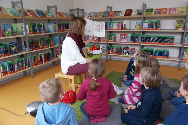 Das Foto wurde im Kinderbereich der Stadtbücherei aufgenommen. Eine Frau sitzt in der Mitte einer Kindergruppe und hält ein Buch zum Vorlesen in der Hand, im Hintergrund sind mehrere Bücherregale zu sehen.