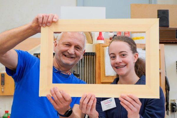 Foto einer erwachsenen männlichen Person auf der linken Seite, sowie jüngeren weiblichen Person auf der rechten Seite. Beide Personen halten einen Holzrahmen in die Höhe und schauen durch den Rahmen hindurch.