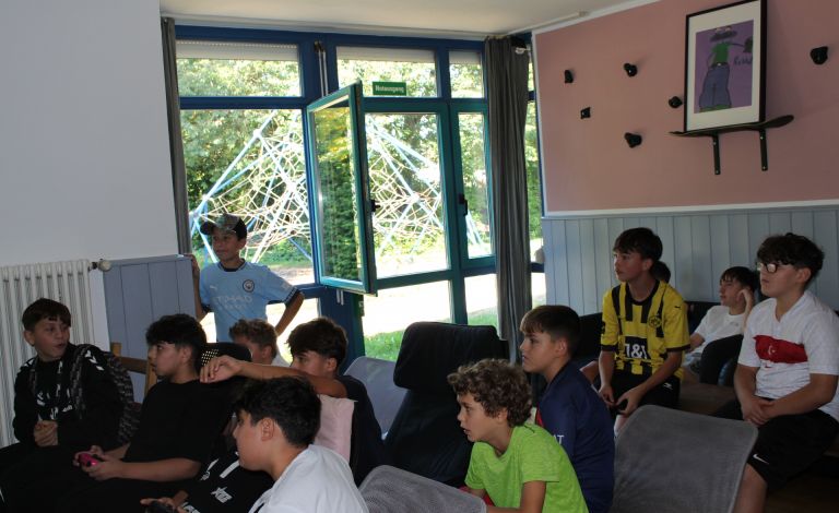 Das Bild wurde in einem Innenraum aufgenommen. Im Hintergrund sieht man ein Fenster. Im Vordergrund sind einige Kinder zu sehen, die gebannt nach links vorne gucken, wo sich der Fernseher befindet, auf dem gerade das Playstation-Spiel Fifa gespielt wird.